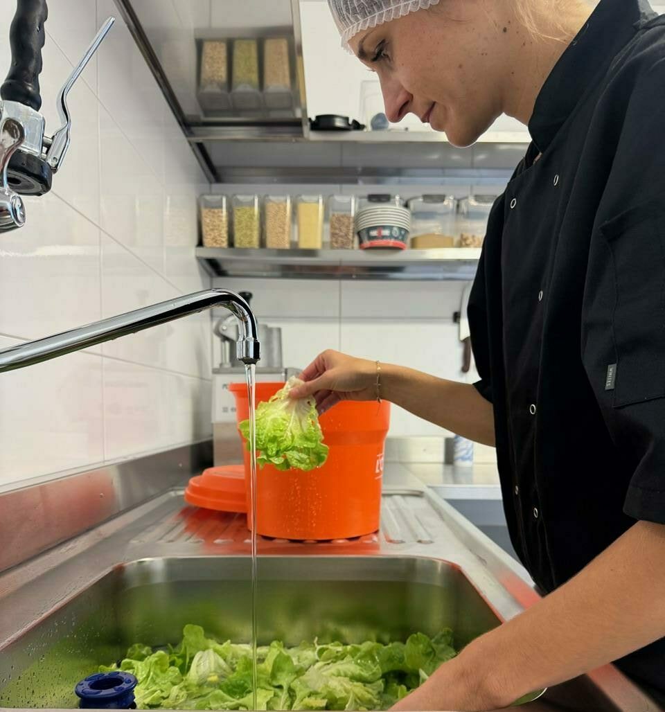 fraich-heure-livraison-de-repas-en-entreprise cuisiniere-habillee-de-noir-lavant-la-salade-poke-bowl-dans-cuisine-industrielle-avec gants-et-charlotte